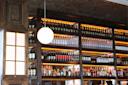 A bar with wooden shelves filled with an assortment of wine and liquor bottles. A round white light fixture hangs in the foreground, and the shelves are softly illuminated, creating a warm, inviting ambiance.