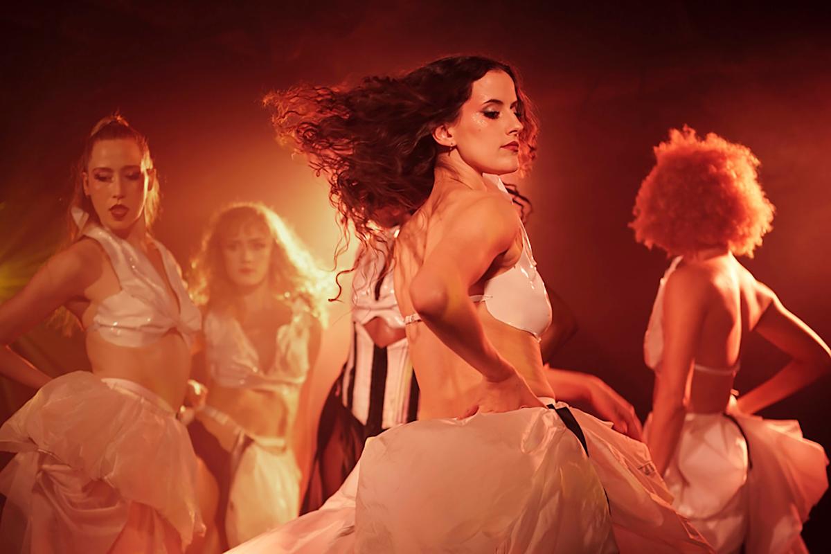 A group of dancers wearing white outfits performs on stage, surrounded by warm, dramatic lighting. The central dancer, with flowing hair, strikes a dynamic pose, conveying energy and movement.