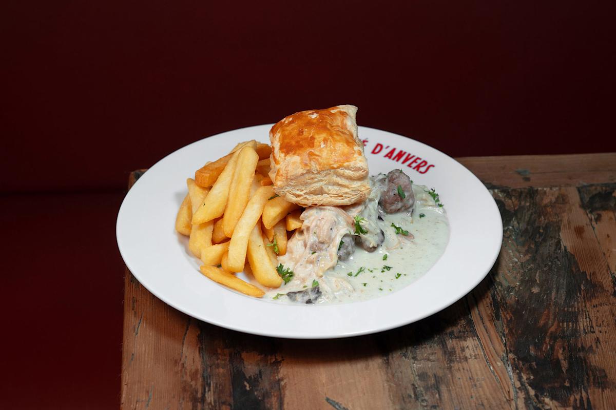 A plate of golden fries, creamy chicken and mushroom stew, and a puff pastry square, served on a white plate with “D’ANVERS” written on the rim, placed on a rustic wooden table against a red background.