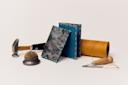 A collection of bookbinding tools including a mallet, awl, needle, thread, and a metal spool. Two books, one with a textured black cover and the other with a smooth blue cover, stand upright against the spool.