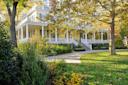A large yellow house with a wraparound porch is surrounded by trees and lush greenery. A curving pathway leads to the front steps. The scene is bathed in sunlight, highlighting the vibrant colors of the foliage.