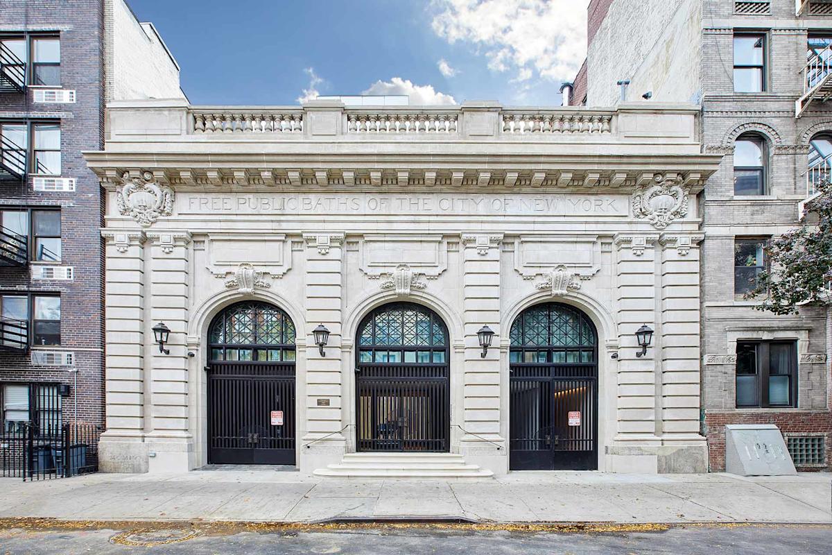 Historic stone building with three large arched doorways, ornate details, and a sign reading "Free Public Baths of the City of New York." Flanked by brick apartment buildings on both sides.