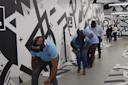 A group of people wearing VR headsets walk in a line along a hallway with geometric black and white wall designs, each person carefully stepping and reaching forward.