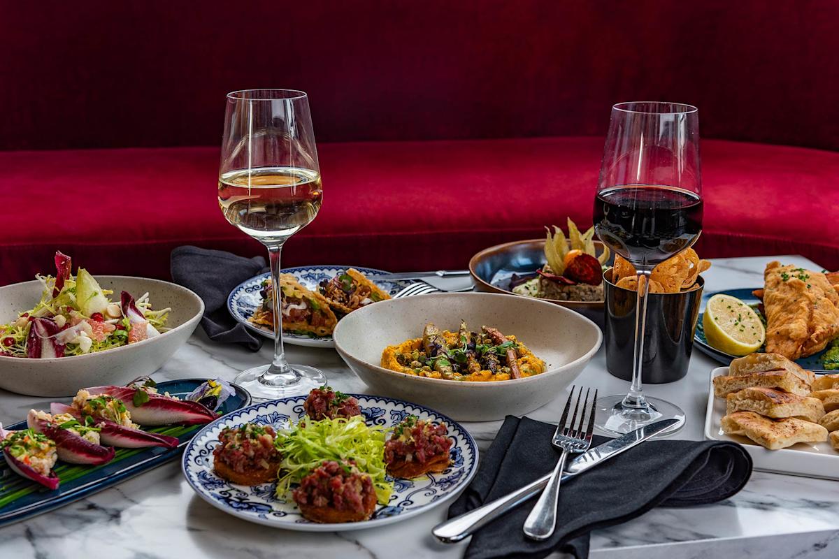 A marble table set with assorted gourmet dishes, including salads, appetizers, small plates, and two glasses of wine—one red and one white—against a red velvet bench background. Cutlery and napkins are also arranged.