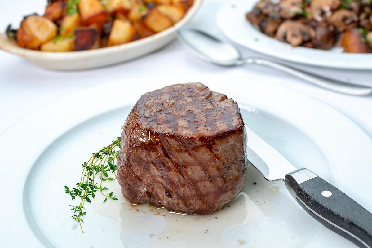 A cooked filet mignon steak sits on a white plate with a sprig of thyme and a steak knife. In the background, plates of roasted potatoes and sautéed mushrooms are visible.