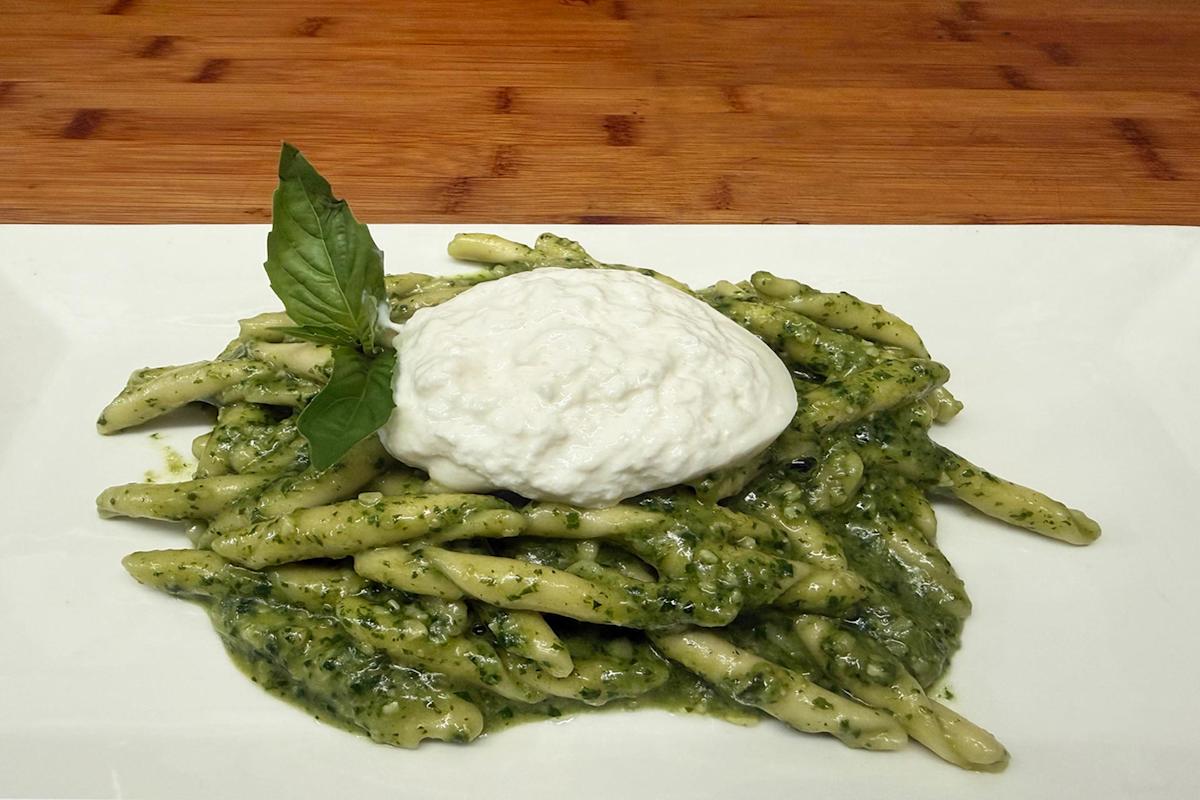 A plate of twisted pasta coated in green pesto sauce, topped with a dollop of creamy white burrata cheese and garnished with a fresh basil leaf, served on a white rectangular dish with a wooden background.