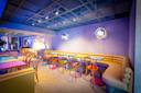 A stylish modern cafe with pastel walls, wooden chairs, colorful table tops, and soft lighting from round wall fixtures. A cushioned bench runs along the wall, creating a cozy, inviting atmosphere.