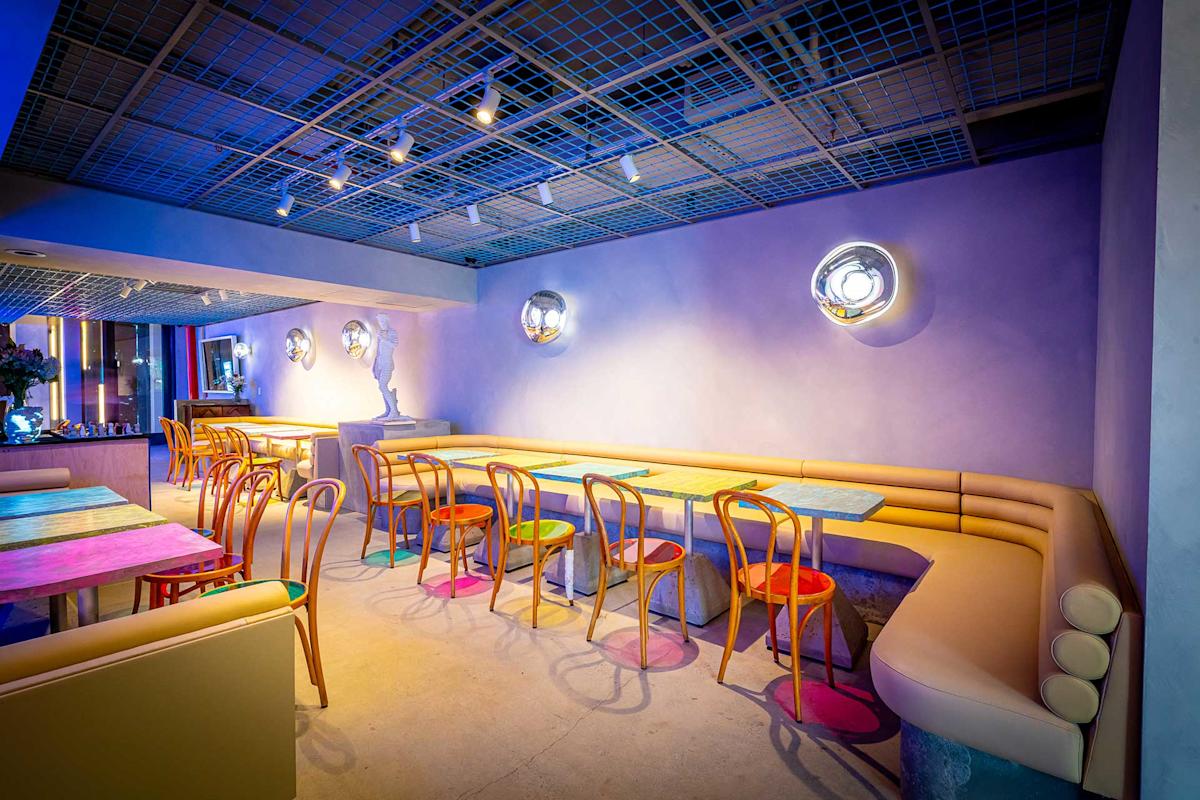 A stylish modern cafe with pastel walls, wooden chairs, colorful table tops, and soft lighting from round wall fixtures. A cushioned bench runs along the wall, creating a cozy, inviting atmosphere.