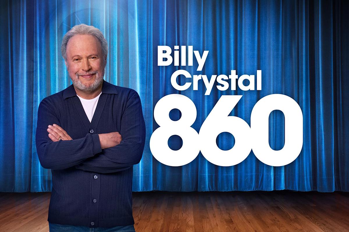 A smiling man with gray hair and beard stands with arms crossed in front of blue stage curtains. Large white text reads: “Billy Crystal 860.”.
