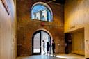 Three people stand at an arched doorway in a brick building, partially open to the outside. Sunlight streams in, and the Brooklyn Bridge is visible through an arched window above the door.