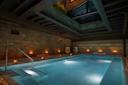 An indoor pool with glowing lights along the brick walls, metal ceiling beams above, and white steps leading into the water, creating a calm and inviting atmosphere.