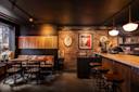 Cozy bar with brick walls, featuring a wooden bar counter and high stools. There are two small tables with chairs by the window. The dimly lit space is decorated with framed art and vintage-style lamps, creating a warm and inviting atmosphere.