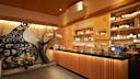 A modern Japanese bar with a black countertop, illuminated wooden slats, shelves stocked with bottles, and a decorative wall featuring a black tree sculpture and floral arrangements. Warm lighting creates a cozy atmosphere.