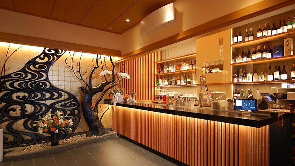 A modern Japanese bar with a black countertop, illuminated wooden slats, shelves stocked with bottles, and a decorative wall featuring a black tree sculpture and floral arrangements. Warm lighting creates a cozy atmosphere.