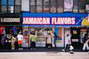 People stand in front of a store called "Jamaican Flavors" with a bright blue and red sign. The sidewalk is busy with pedestrians, and colorful posters and clothing are displayed outside nearby shops.