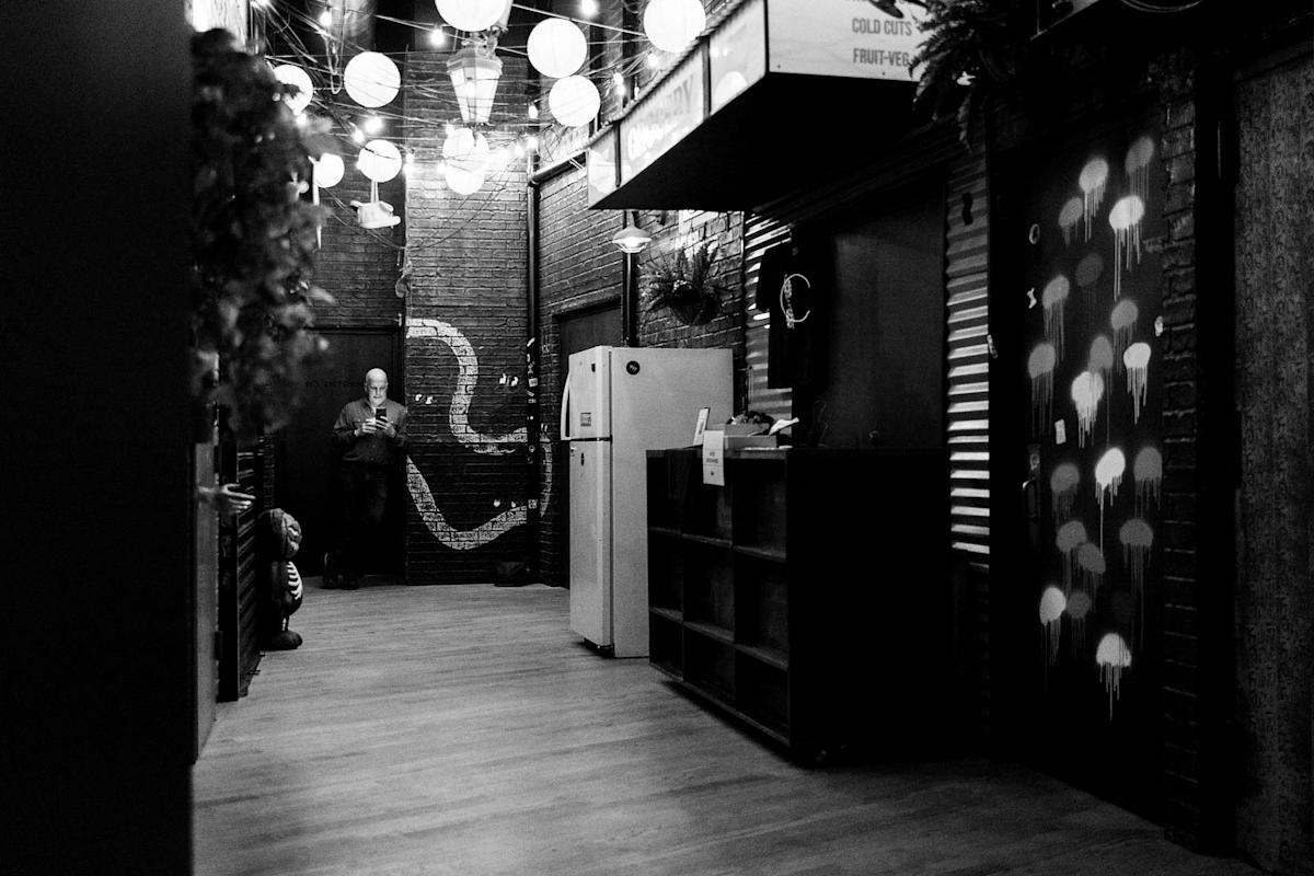 A grayscale photo of a dimly lit hallway adorned with hanging lanterns. A person stands in the back, looking at a phone. Walls feature artwork; one side decorated with abstract patterns, the other with glowing jellyfish graffiti.