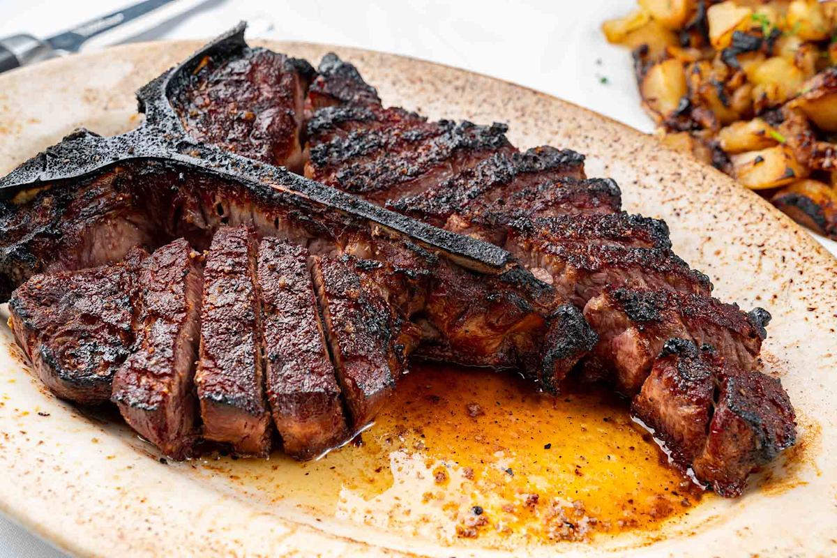 A sliced, juicy, grilled T-bone steak on a speckled plate with juices pooling around it, served alongside a portion of roasted potatoes.