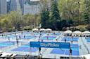 Multiple people play pickleball on several blue outdoor courts surrounded by trees in a city park, with tall buildings visible in the background. A banner reads "CityPickle" in the foreground.