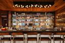 A modern bar with stylish wooden decor featuring a variety of liquor bottles on shelves behind the counter. Three wooden bar stools are in front, and a unique ceiling light fixture adds ambiance