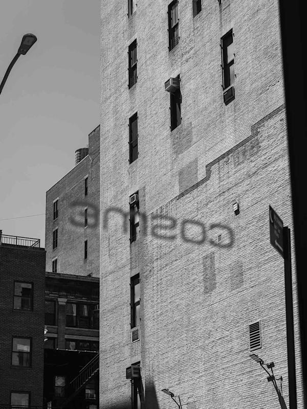 A black and white photo of tall brick buildings. The shadow of a sign reading "Cosine" is cast on one of the buildings. A streetlamp and portions of fire escapes are also visible.