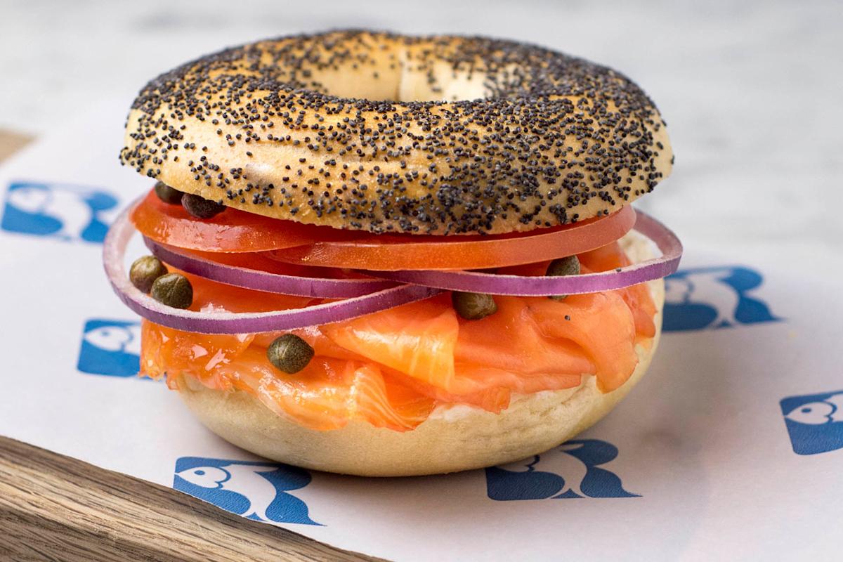 A poppy seed bagel sandwich with layers of smoked salmon, sliced tomatoes, red onions, and capers, served on a sheet of branded paper.