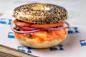 A poppy seed bagel sandwich with layers of smoked salmon, sliced tomatoes, red onions, and capers, served on a sheet of branded paper.