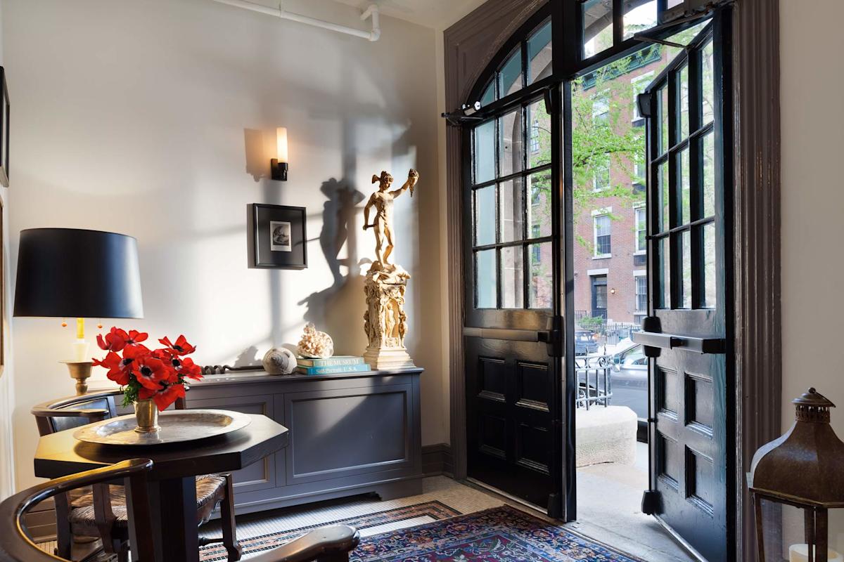 A bright entryway with an open black door, a side table with red flowers, a black lamp, a sculpture on a cabinet, a wall sconce, framed art, and a view of trees and brick buildings outside.