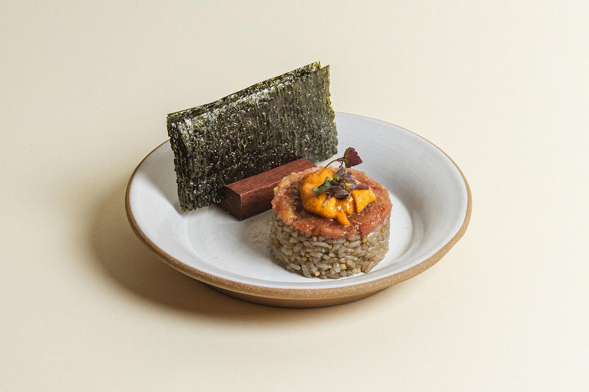 A plated dish featuring a round serving of rice topped with orange sea urchin and garnish, accompanied by a rectangular cracker and a sheet of nori, served on a white plate with a tan rim.