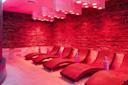 A modern spa relaxation room with several curved lounge chairs and textured stone walls, illuminated by ambient pink-red lighting from ceiling fixtures.