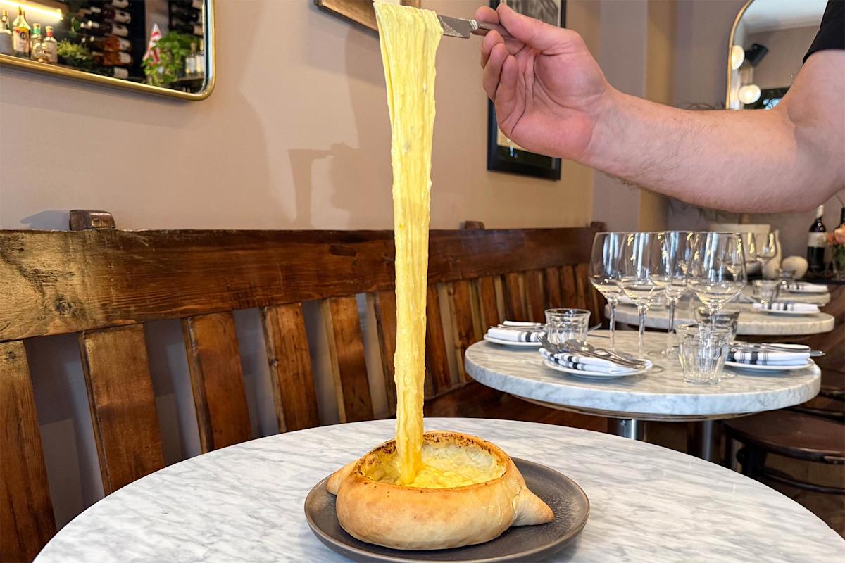 A hand lifts a knife with stretchy melted cheese from a boat-shaped bread filled with cheese, set on a small plate atop a marble table in a cozy restaurant setting.