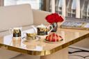 A polished table set with a gold-accented tea set, a cup of tea, a plate with a croissant, a vase of red roses, and a book titled "Alan Faena," in front of a beige sofa and large windows.