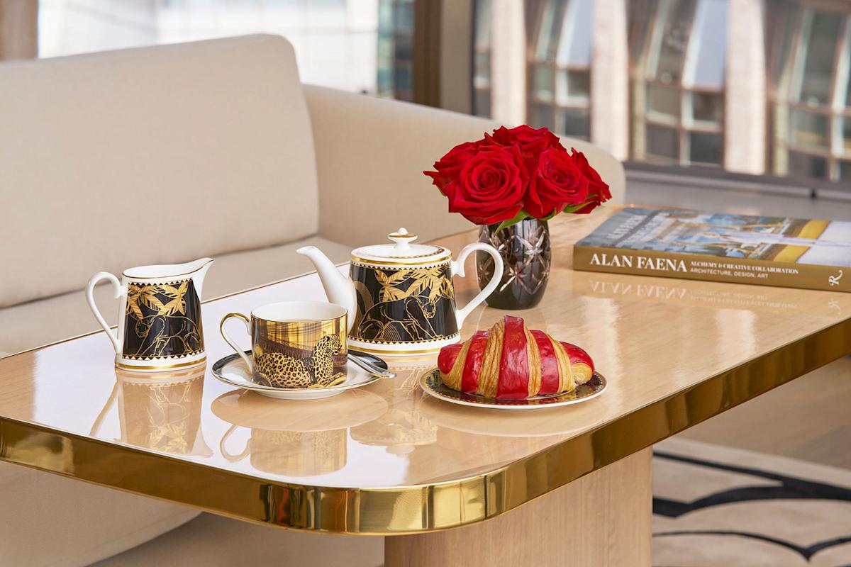 A polished table set with a gold-accented tea set, a cup of tea, a plate with a croissant, a vase of red roses, and a book titled "Alan Faena," in front of a beige sofa and large windows.