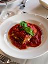 A slice of lasagna topped with tomato sauce and fresh basil leaves is served on a white plate, with a fork, knife, and napkin visible on the table in the background.