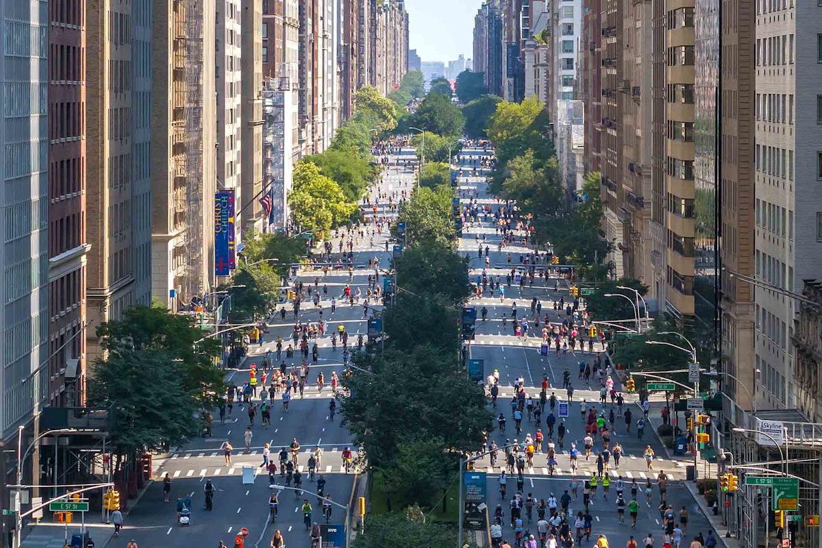 A city street closed to traffic, filled with people walking and cycling between tall buildings under a clear sky, with trees lining the sidewalks.