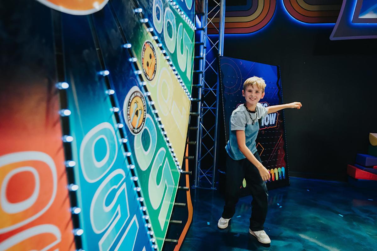 A smiling child throws a ball at a brightly colored arcade game in a neon-lit room, aiming to hit targets on the game board.