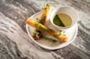 Two crispy rolled appetizers garnished with green herbs are served on a white plate beside a small bowl of light green dipping sauce, all placed on a marbled surface.