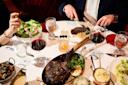 A table set for two with a large tomahawk steak, salads, glasses of red wine, water, and whiskey. One person is cutting steak while the other holds a fork. Sides and sauces are also present on a white tablecloth.
