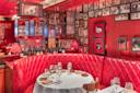 A cozy restaurant features a red leather tufted booth, a round table set with wine glasses and plates, warmly lit by red lamps, with walls covered in framed black-and-white photos and patterned red wallpaper.