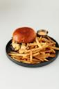A cheeseburger with a shiny bun and melted cheese sits on a black plate, served with a large portion of golden French fries and a small cup of dipping sauce.