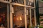 The image shows the exterior of Antica Pesa restaurant, with glass windows revealing a warmly lit interior, tables set for dining, and the restaurant’s name and logo visible on the inside wall.