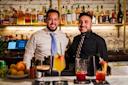 Two smiling bartenders stand behind a bar with a variety of colorful cocktails in front of them. Bottles of liquor are displayed on shelves against a white brick wall in the background.