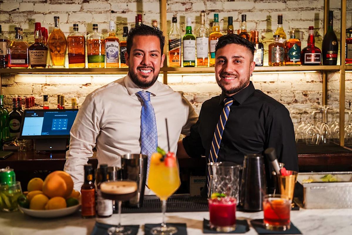 Two smiling bartenders stand behind a bar with a variety of colorful cocktails in front of them. Bottles of liquor are displayed on shelves against a white brick wall in the background.