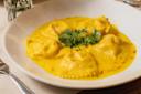A white plate with five ravioli pieces in a creamy yellow saffron sauce, garnished with fresh parsley in the center. A fork and a glass are visible at the edges of the image.