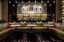 A modern, stylish lounge with curved beige sofas, round tables, black ottomans, and colorful abstract artwork on the wall above a well-lit bar stocked with bottles and glassware. Warm, ambient lighting hangs from the ceiling.