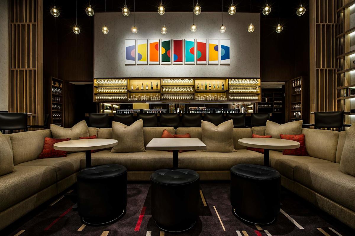 A modern, stylish lounge with curved beige sofas, round tables, black ottomans, and colorful abstract artwork on the wall above a well-lit bar stocked with bottles and glassware. Warm, ambient lighting hangs from the ceiling.