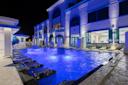 A luxurious outdoor pool at night, illuminated with blue lights, features lounge chairs partially in the water and a grand, elegant building in the background.