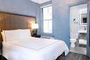 A modern hotel room with a neatly made bed, bedside lamp, large window, and an open door showing a bathroom with a toilet, sink, and well-lit mirror. The walls are painted light blue.