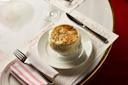 A bowl of French onion soup topped with melted, browned cheese sits on a white plate with a doily, beside a knife and a fork wrapped in a striped napkin on a restaurant table.