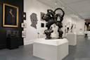 Art gallery interior featuring sculptures, paintings, and photographs. Central focus is on a bronze sculpture of two heads with intricate hair designs. Walls are adorned with various framed artworks, and the space is well-lit and modern.