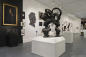 Art gallery interior featuring sculptures, paintings, and photographs. Central focus is on a bronze sculpture of two heads with intricate hair designs. Walls are adorned with various framed artworks, and the space is well-lit and modern.